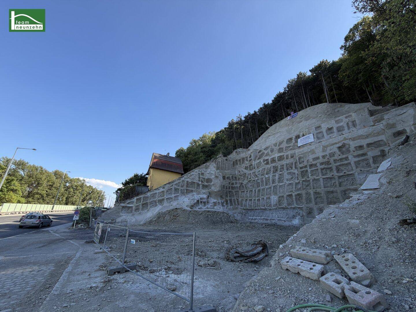 Weitläufige Ansicht eines Hanggrundstücks mit gelbem Haus, großer Stützmauer und angrenzender Straße.