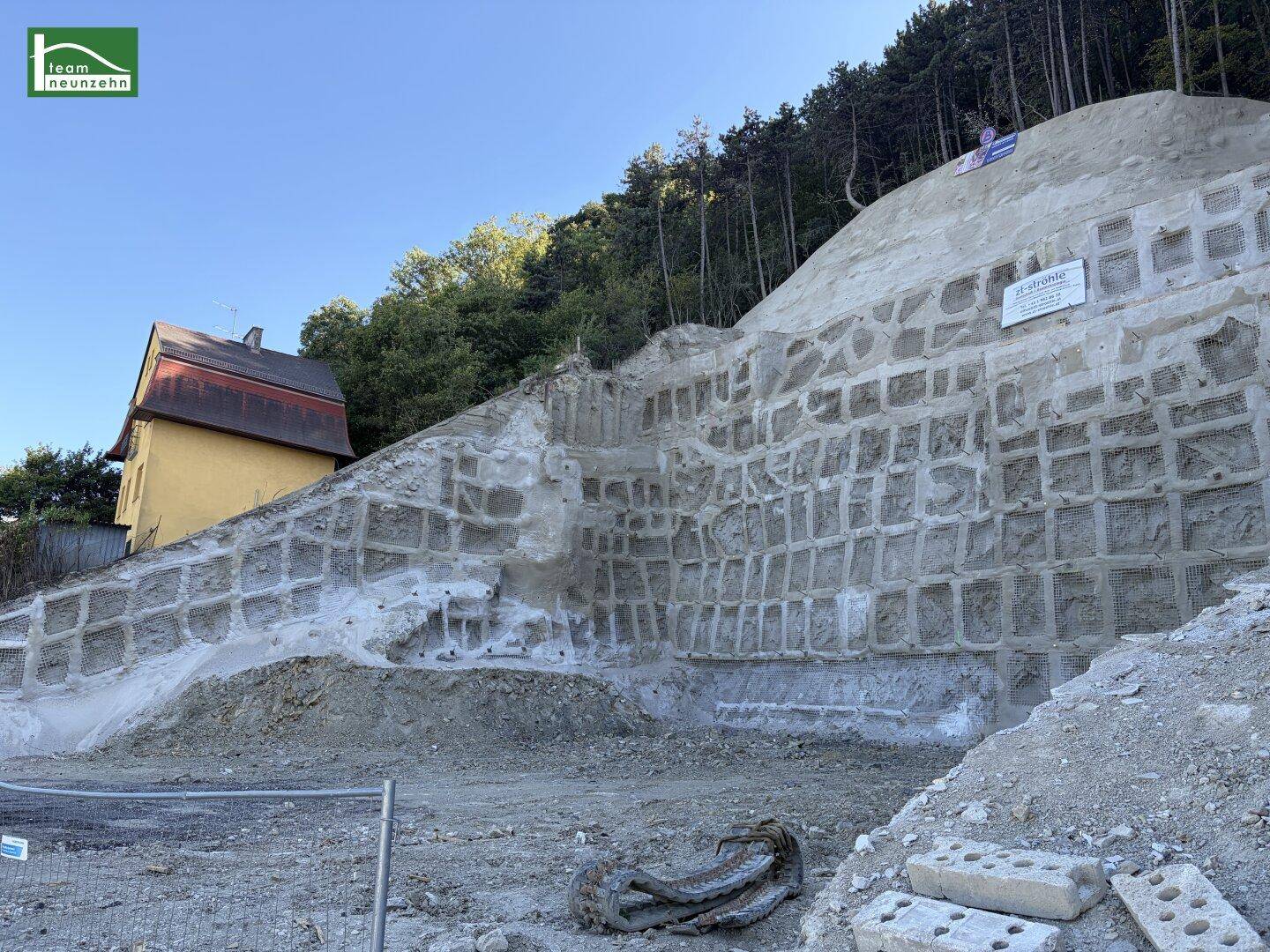 Detailansicht der massiven Stützmauer am Hang mit dem gelben Haus im Hintergrund und Wald.