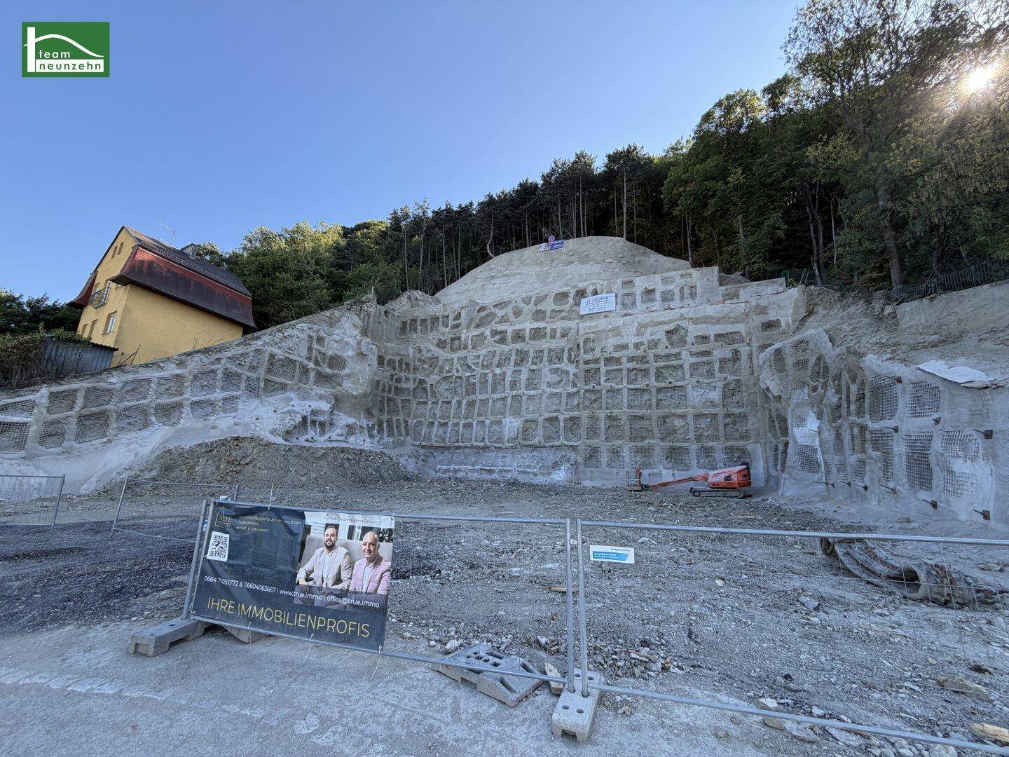 Panoramablick auf das Hanggrundstück mit Stützmauer, gelbem Haus und einem Werbebanner im Vordergrund.