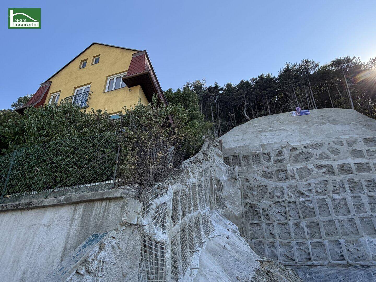 Gelbes Haus auf einem Hanggrundstück mit einer großen, teilweise fertiggestellten Stützmauer und Wald im Hintergrund.