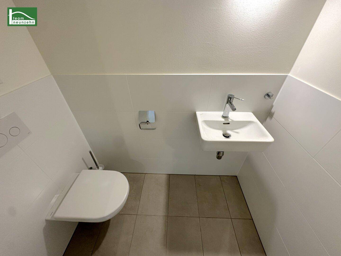 Separates WC mit wandhängender Toilette, kleinem Waschbecken und hellen Fliesen.
