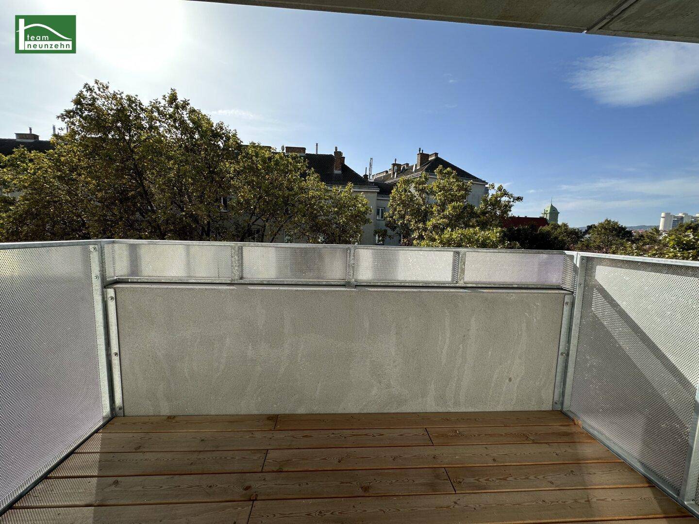 Balkon mit Holzboden und Blick auf die umliegenden Gebäude und grüne Bäume.