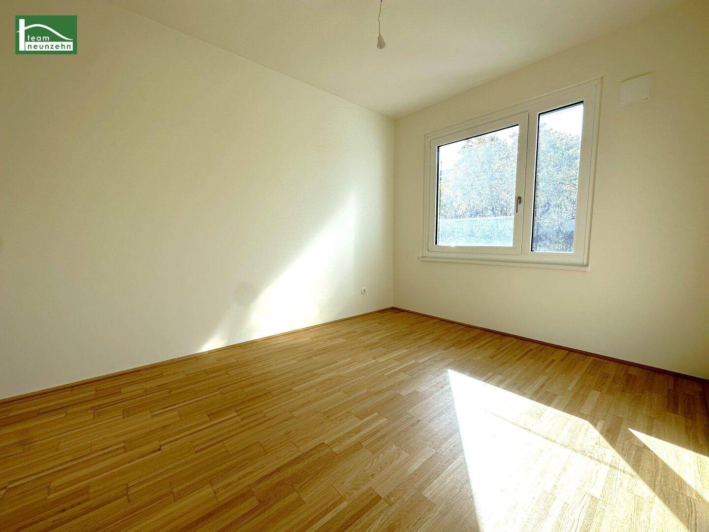 Helles, leeres Schlafzimmer mit Parkettboden und großem Fenster, das viel Tageslicht hereinlässt.