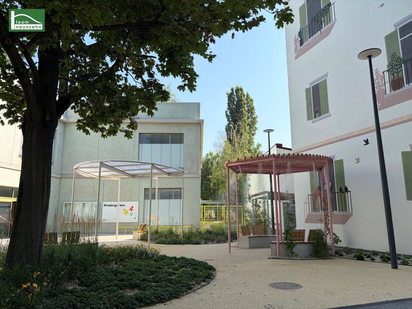 Außenbereich eines Kindergartens mit modernen Gebäuden, Spielplatzstrukturen und grünen Bäumen unter blauem Himmel.