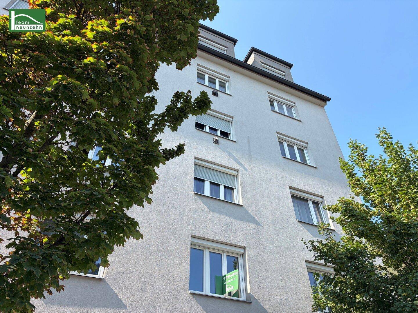 Mehrstöckiges Wohngebäude mit hellgrauer Fassade, vielen Fenstern und Rollläden, umgeben von grünen Bäumen.