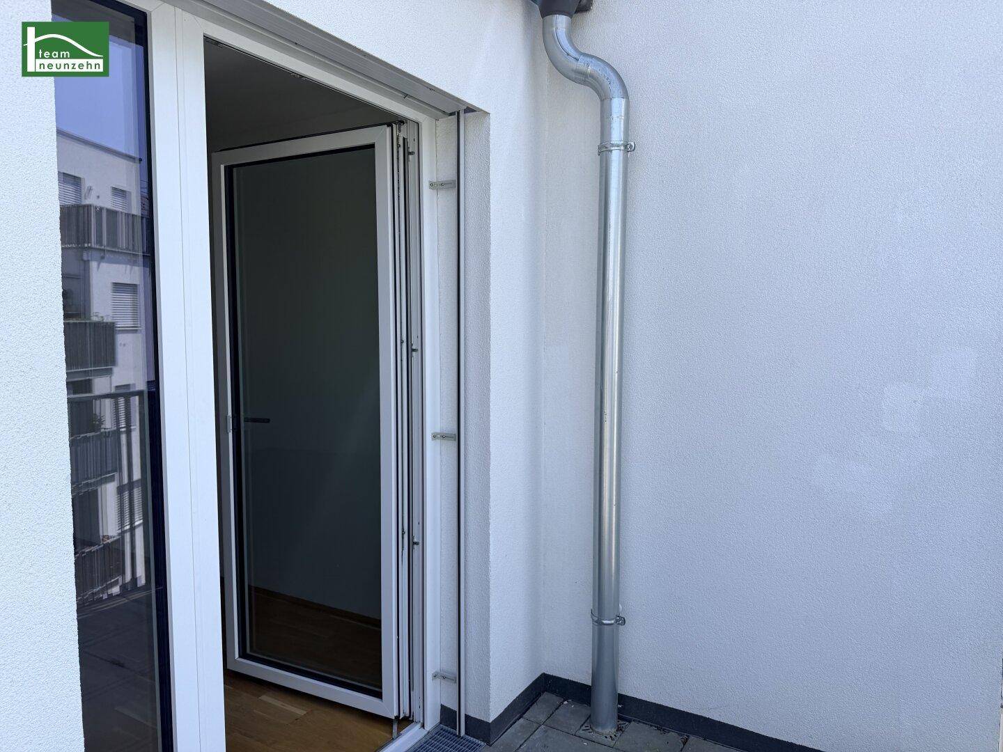 Offene Balkontür führt zu einem Außenbereich mit Regenfallrohr an der weißen Fassade.