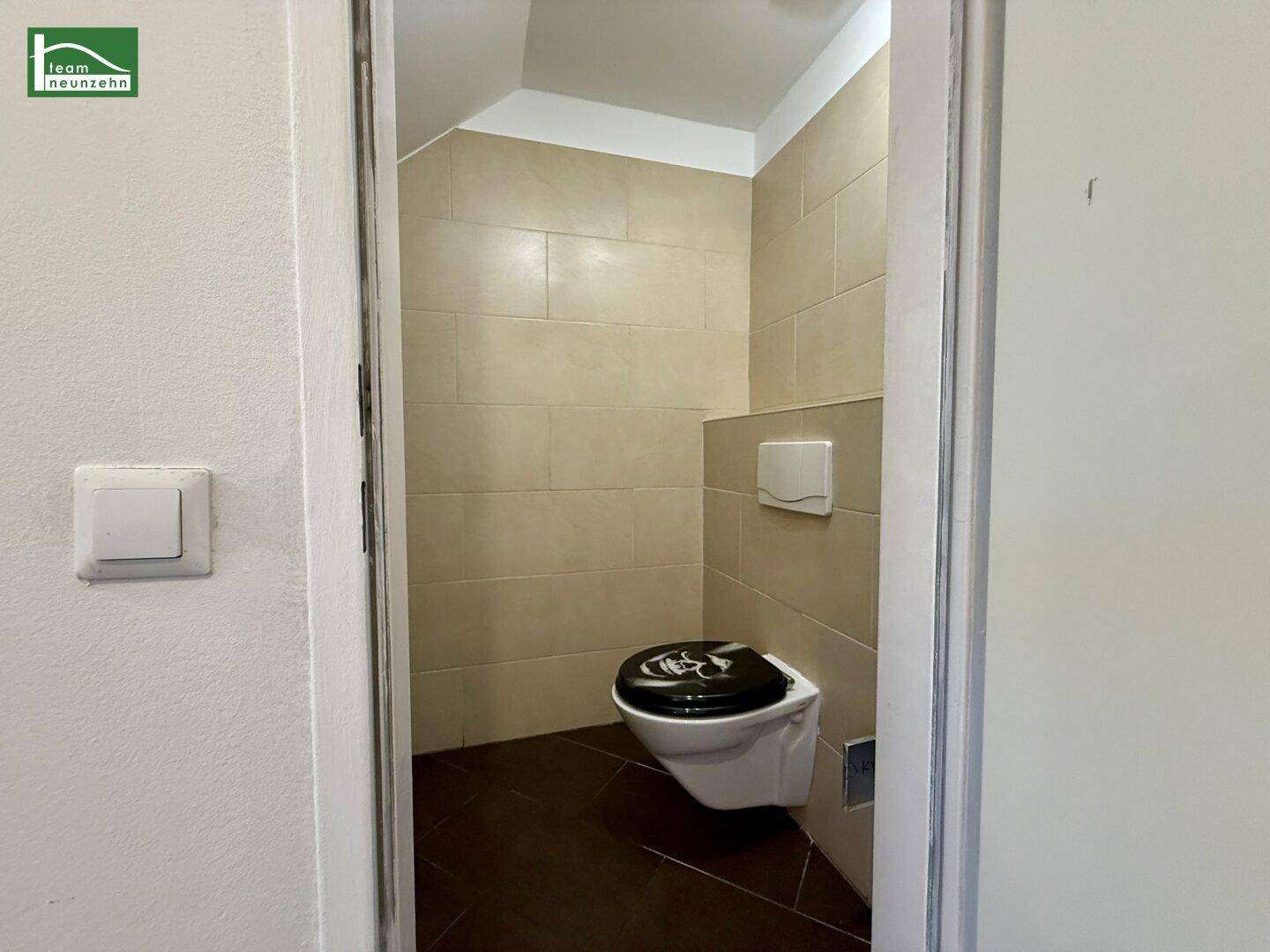 Kleines Gäste-WC mit beigefarbenen Fliesen, dunklem Boden und wandhängendem WC.