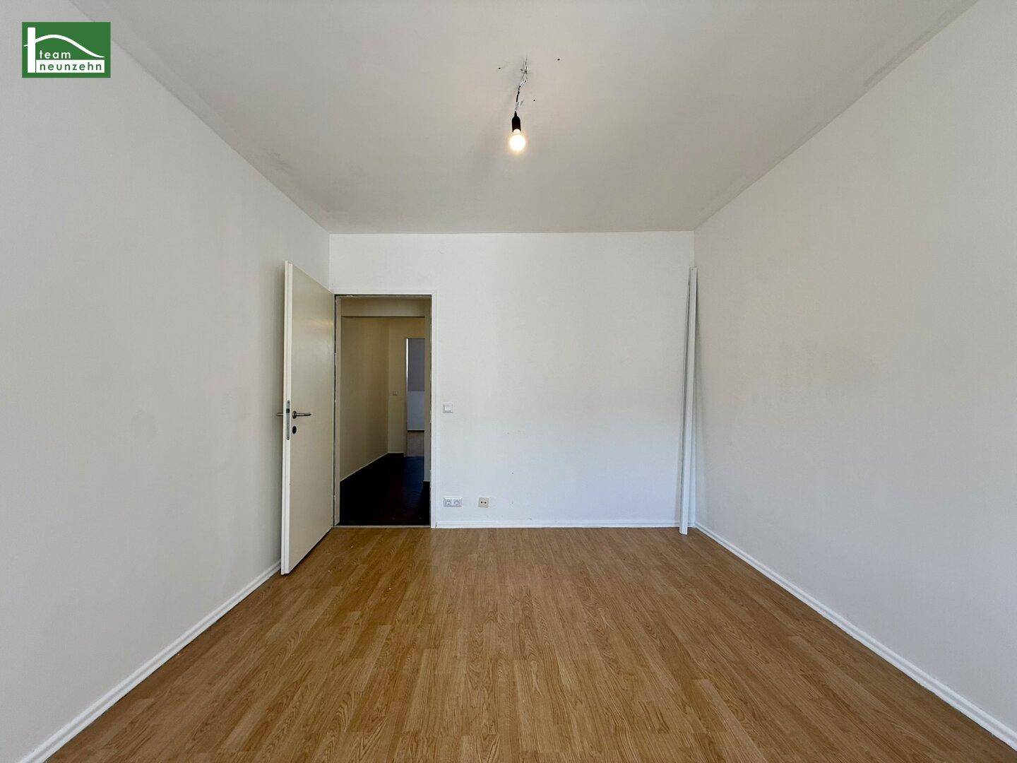 Leerer Raum mit hellem Holzfußboden und weiß gestrichenen Wänden, mit Zugang zu einem Flur.