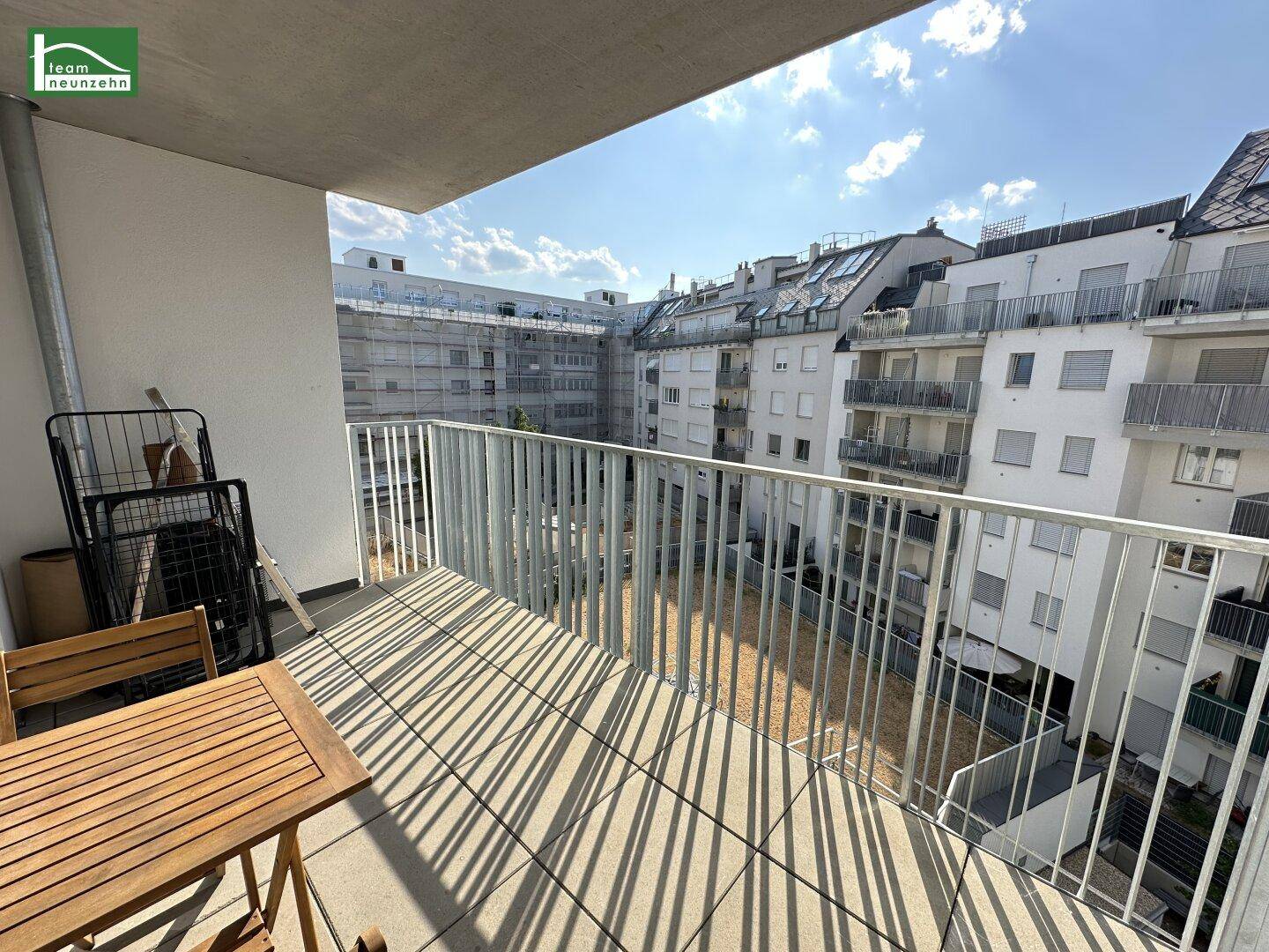 Sonniger Balkon mit Holztisch und Blick auf den Innenhof der Wohnanlage.