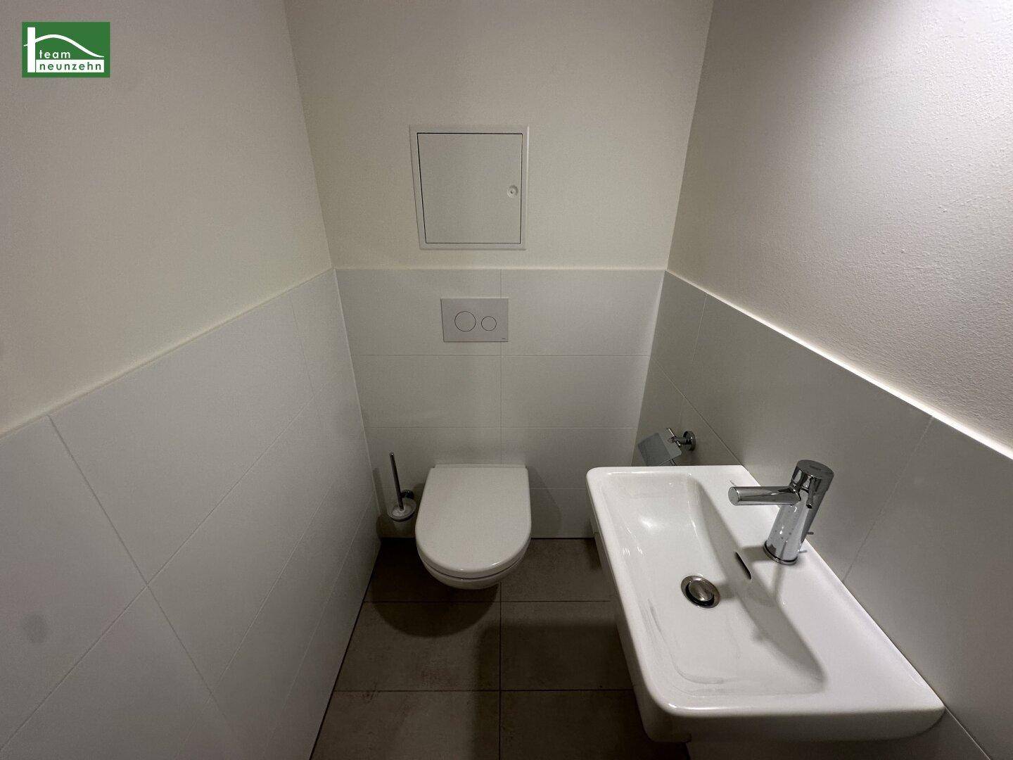 Separates Gäste-WC mit Wand-WC und kleinem Handwaschbecken, halbhoch gefliest in Weiß.