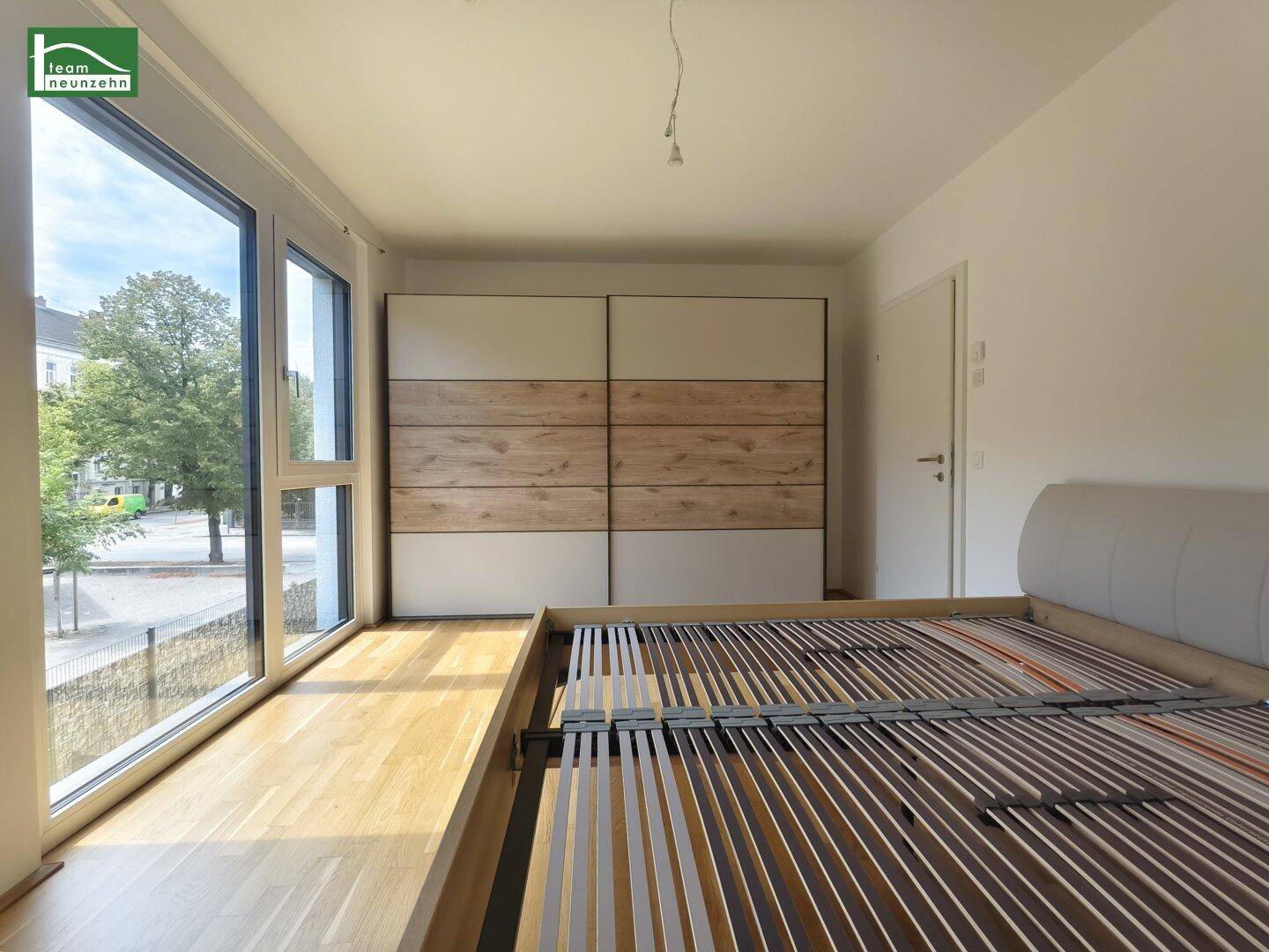 Schlafzimmer mit großen Fenstern, Holzboden und einem Einbauschrank.