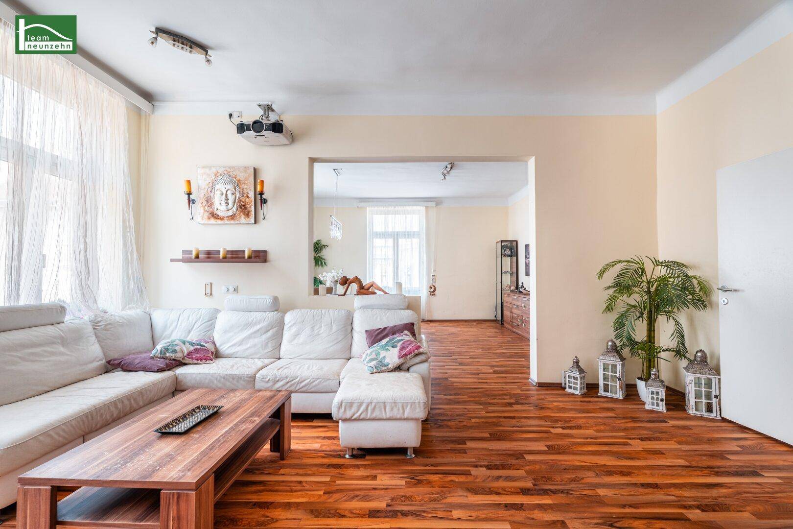 Geräumiges Wohnzimmer mit beigem Sofa, Holzboden und einem Projektor für Unterhaltung.