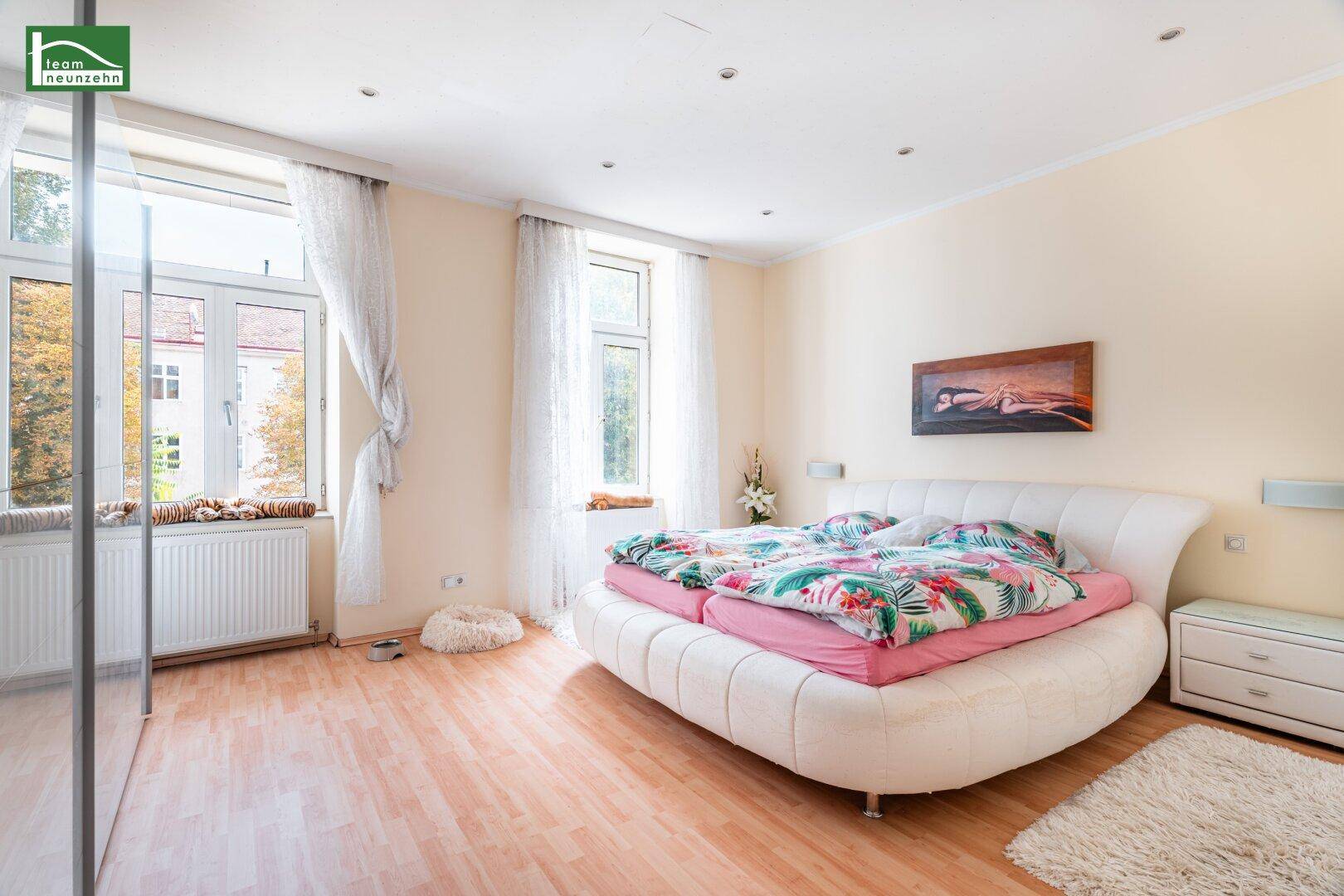 Helles Schlafzimmer mit Doppelbett, Laminatboden und großen Fenstern.
