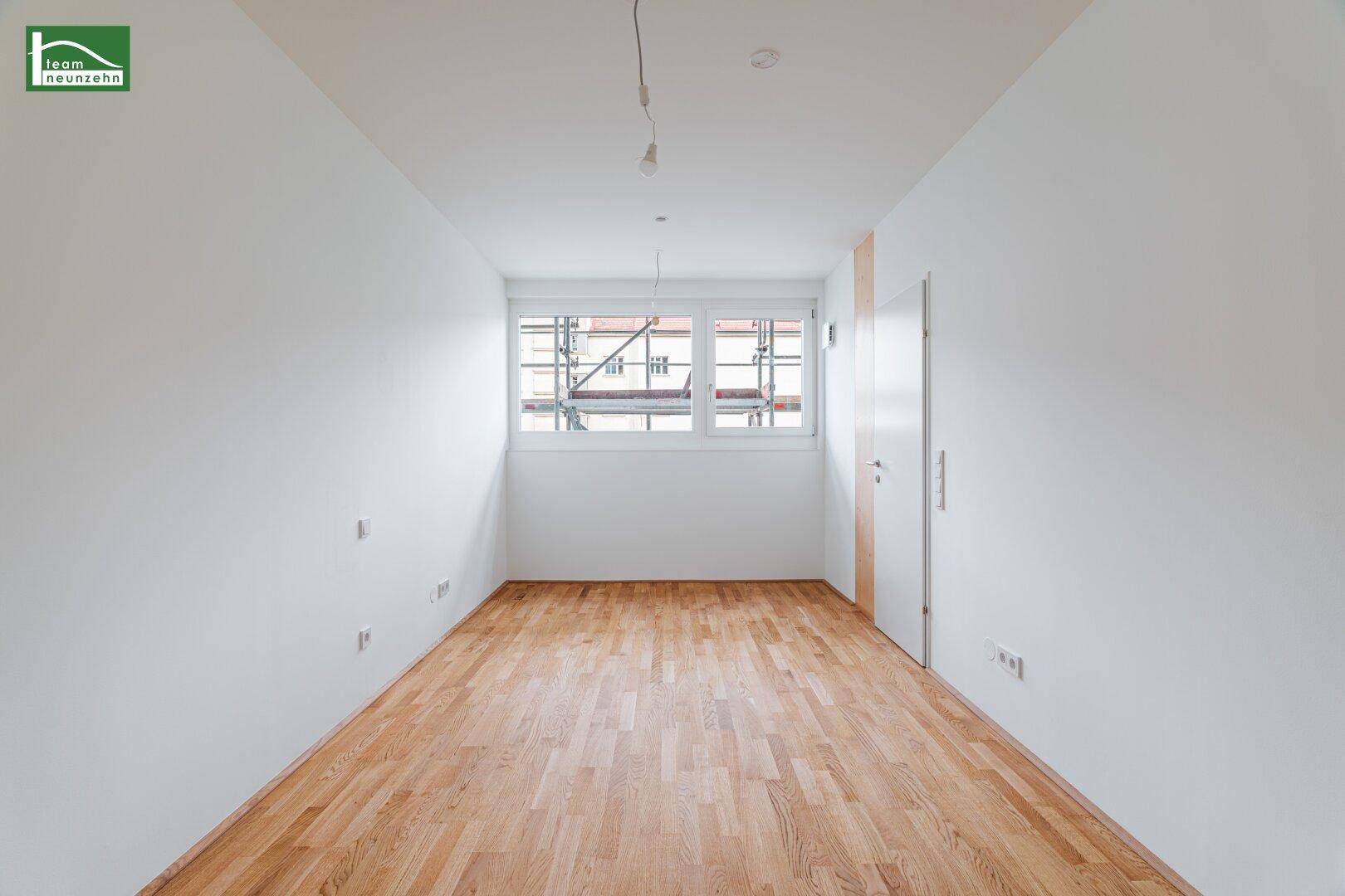 Leeres Zimmer mit Holzboden und Fenster, das einen Blick auf eine Baustelle bietet.
