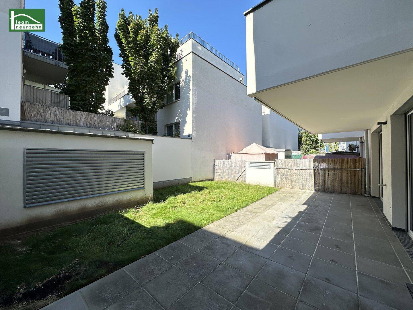 Sonniger Gartenbereich mit gepflasterter Terrasse und Rasenfläche, umgeben von einem Zaun.