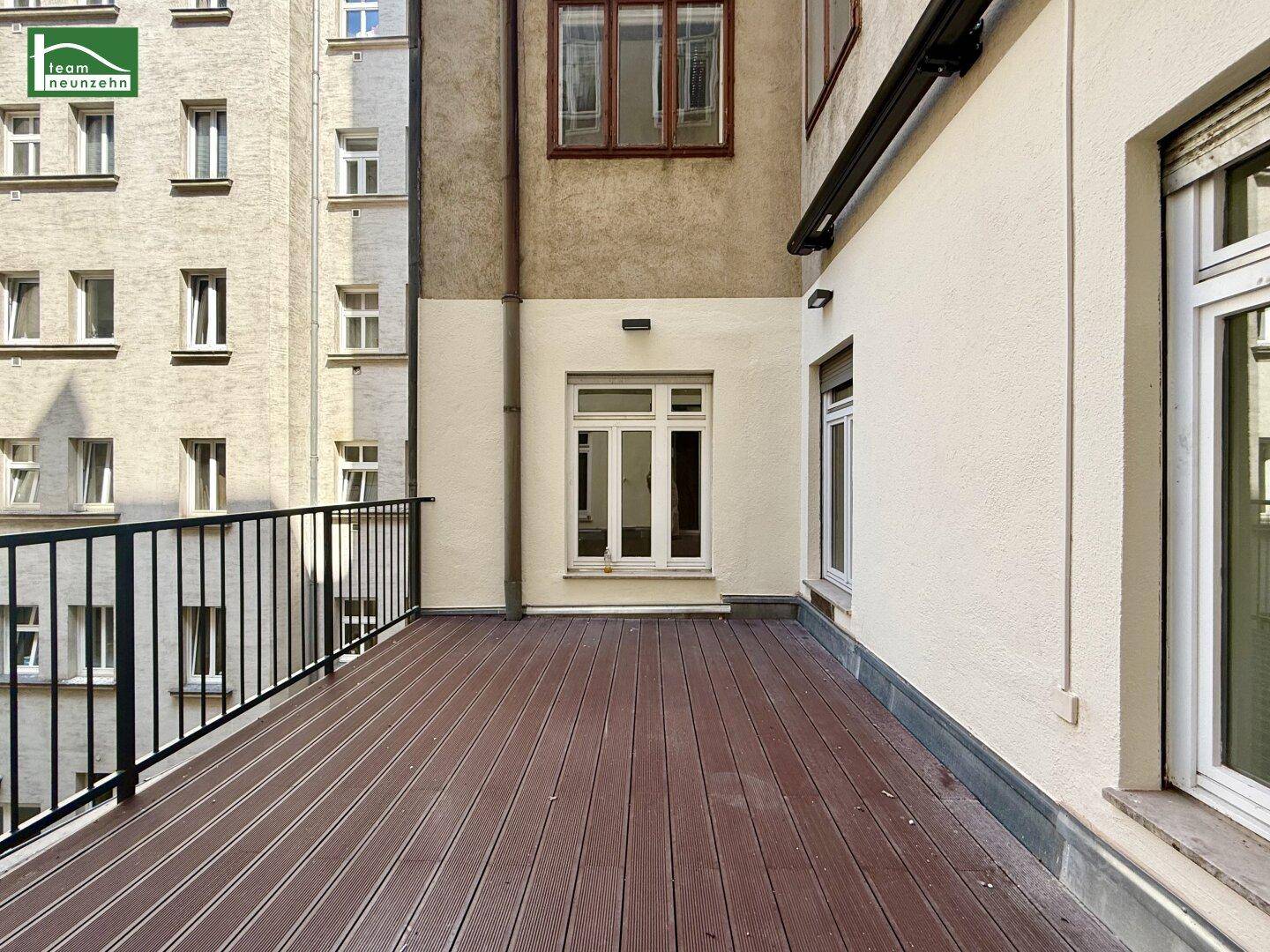 Geräumige Terrasse mit Holzplankenboden und schwarzem Geländer, umgeben von Gebäudefassaden.