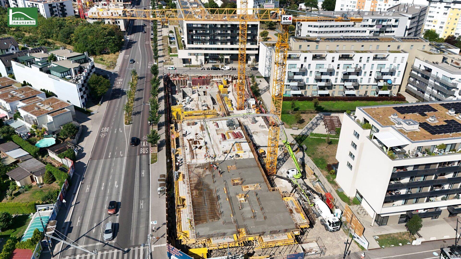 Luftaufnahme einer aktiven Baustelle mit Betonarbeiten und Kränen, umgeben von städtischer Infrastruktur.