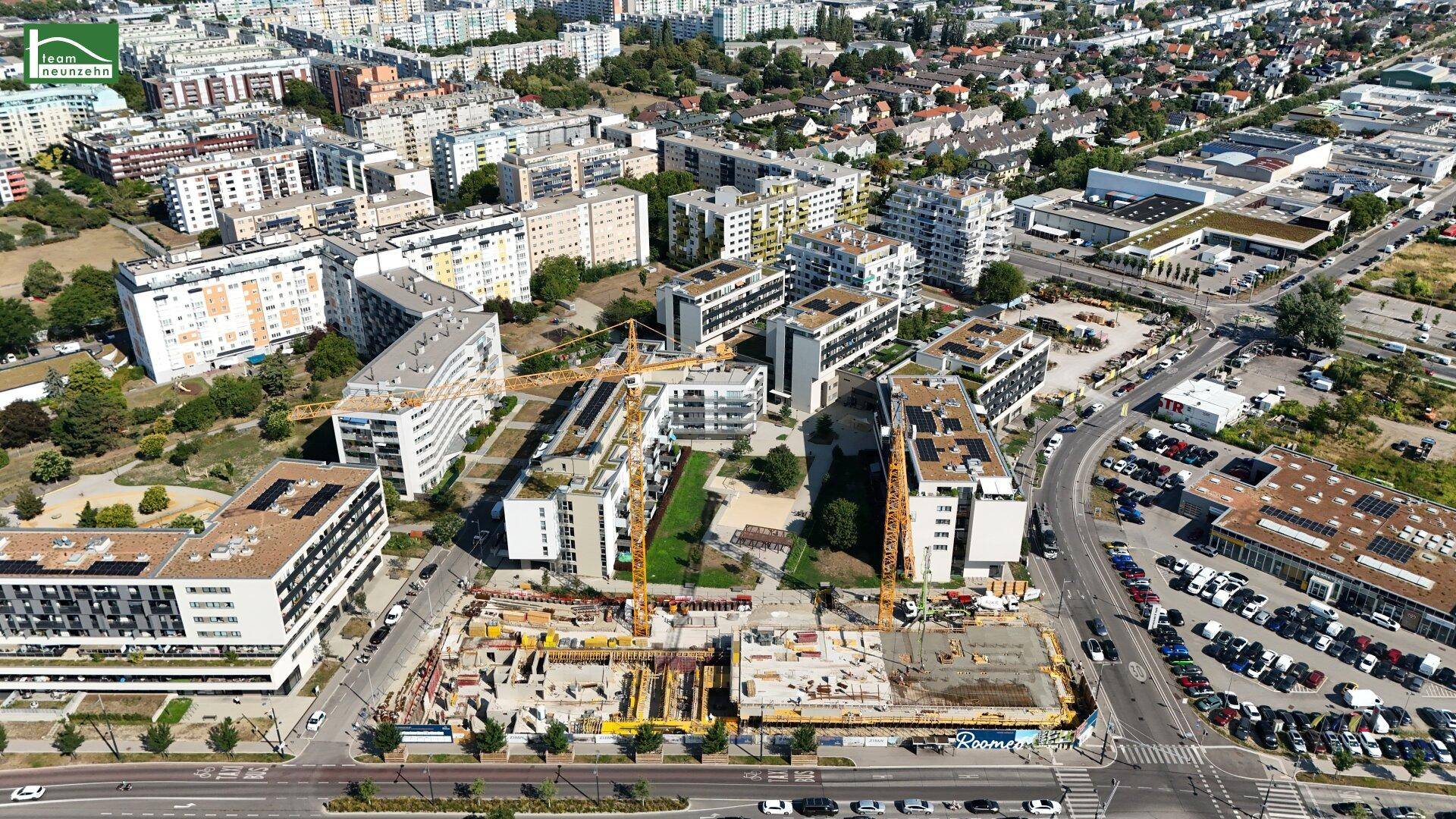 Weitläufige Luftaufnahme einer Baustelle in einem dicht besiedelten Stadtgebiet mit vielen Wohnkomplexen und Grünflächen.