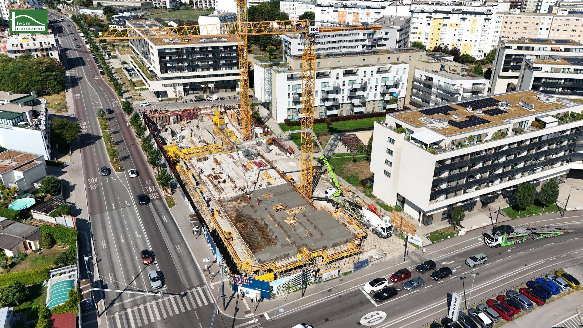 Luftbild der Baustelle mit Kränen und angrenzender Hauptstraße, umgeben von modernen Wohngebäuden und Parkplätzen.