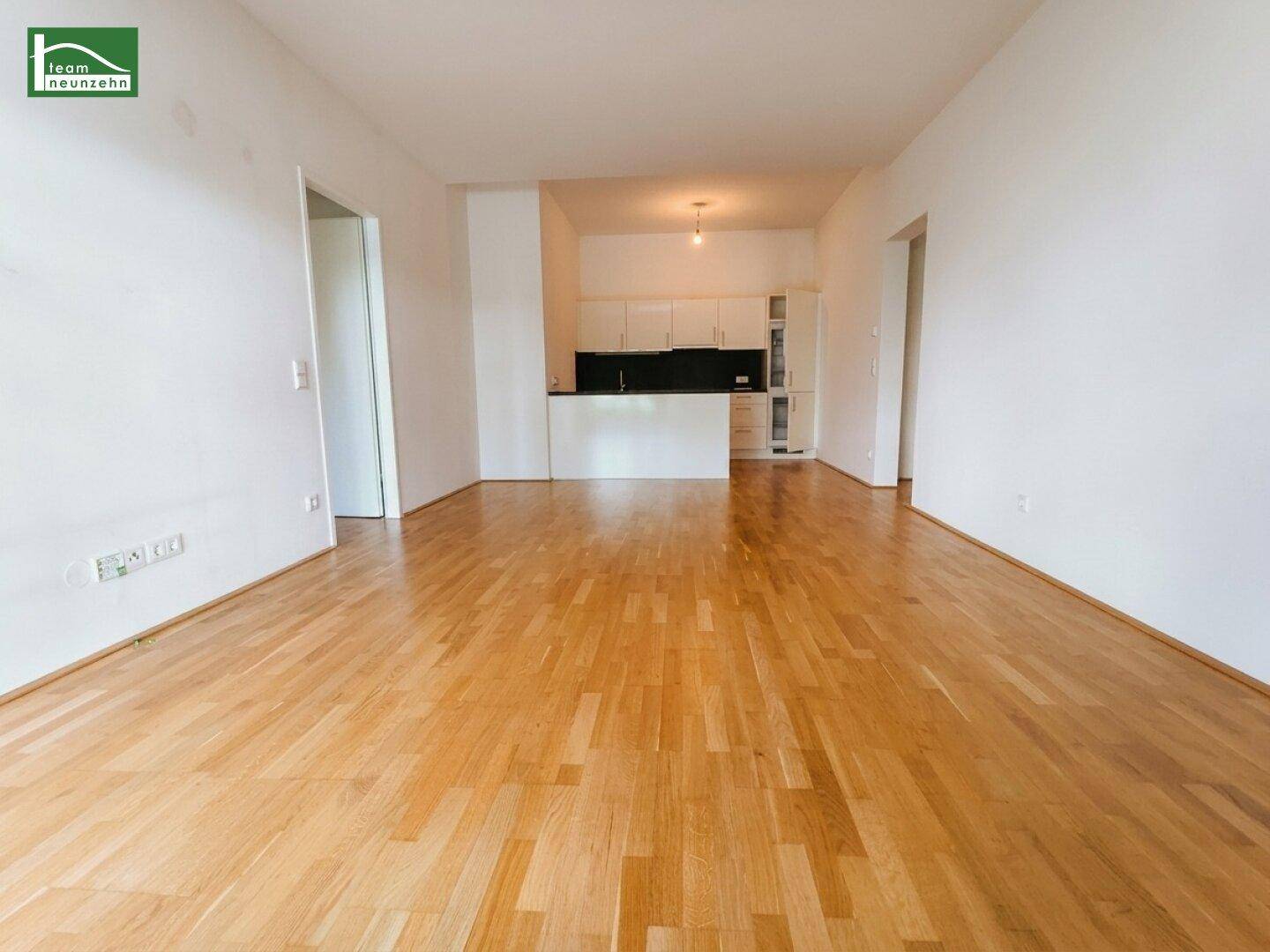 Leerer Wohnbereich mit Holzboden und integrierter Küchenzeile im Hintergrund.