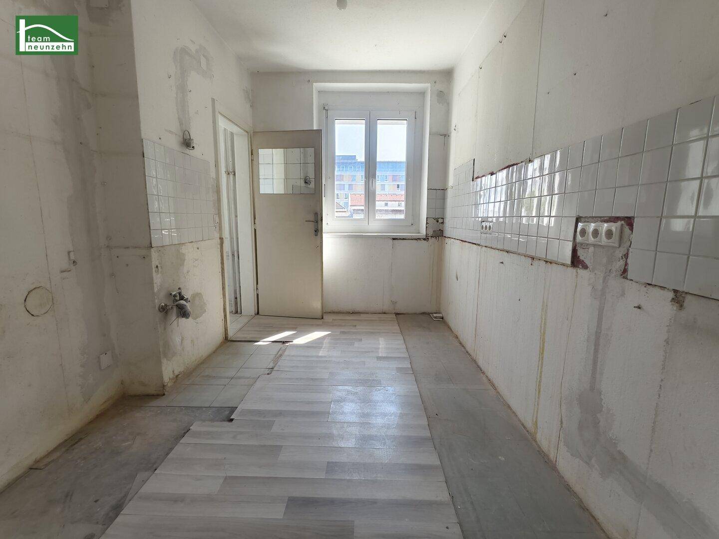 Leere Küche mit weißen Wandfliesen, Fenster und vorbereiteten Anschlüssen für Geräte.