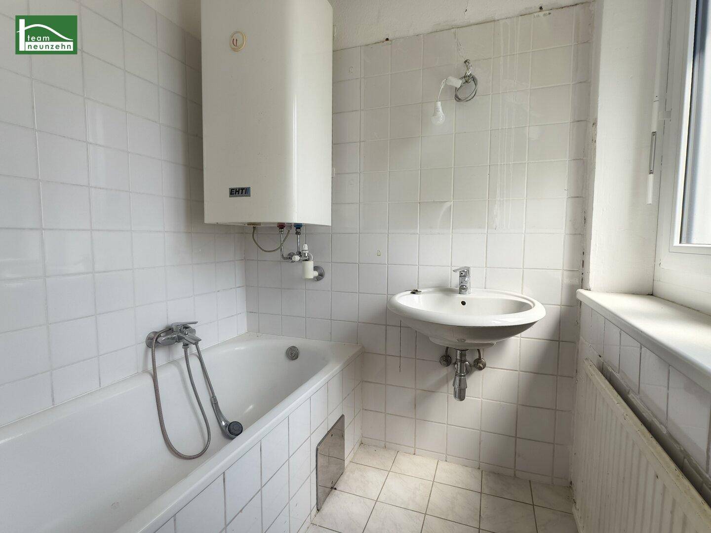 Badezimmer mit Badewanne, Waschbecken, Boiler und weißen Wand- und Bodenfliesen.