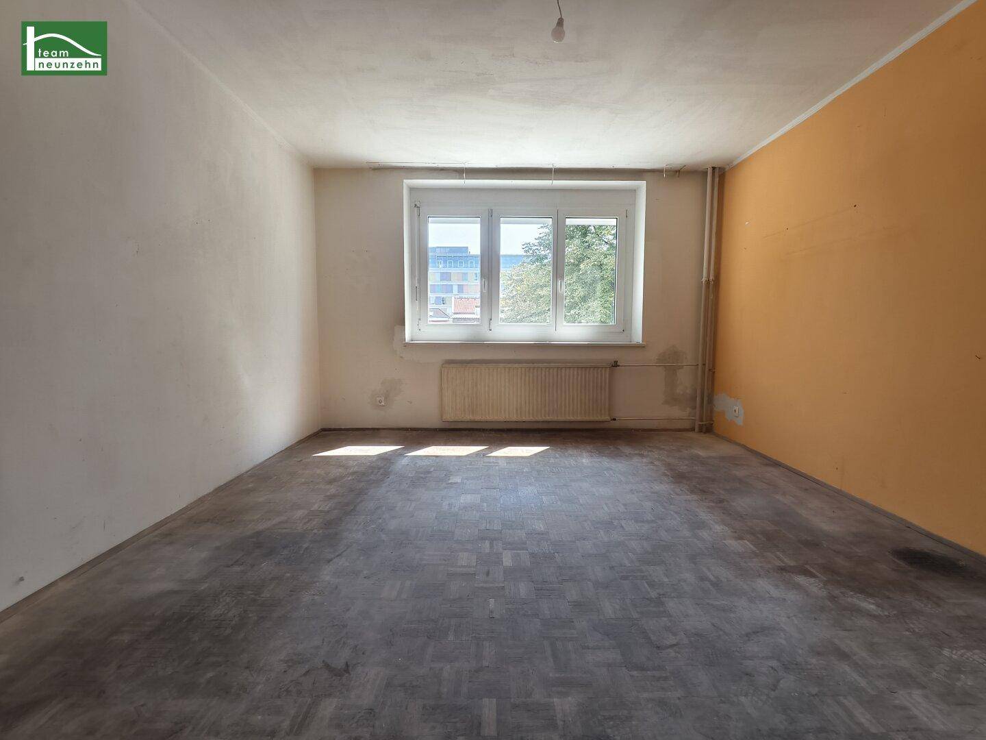 Leeres Zimmer mit abgenutztem Parkettboden, gelber Wand und Fenster.