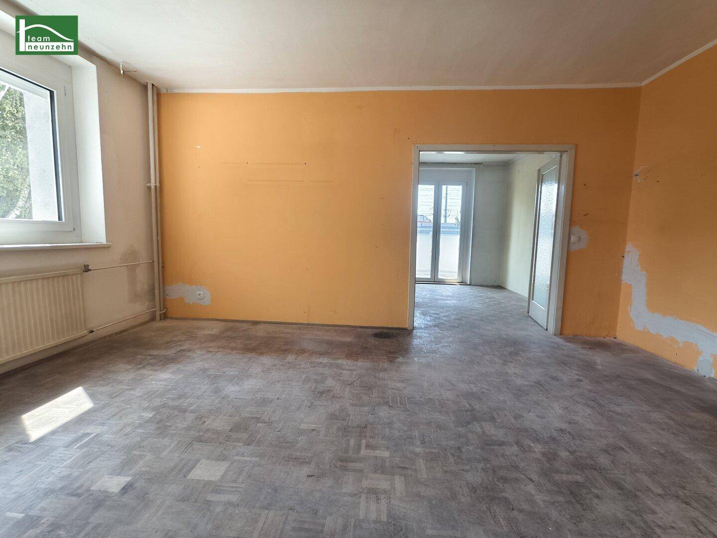 Geräumiges Zimmer mit abgenutztem Parkettboden, gelber Wand und Durchgang zu einem weiteren Raum.