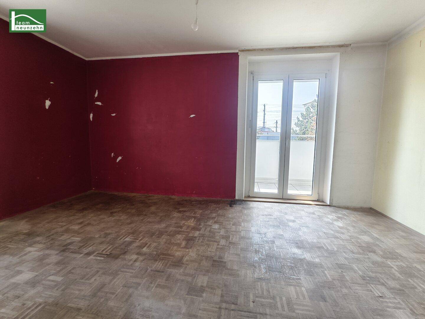 Geräumiges Zimmer mit abgenutztem Parkettboden, roter Akzentwand und Zugang zum Balkon.