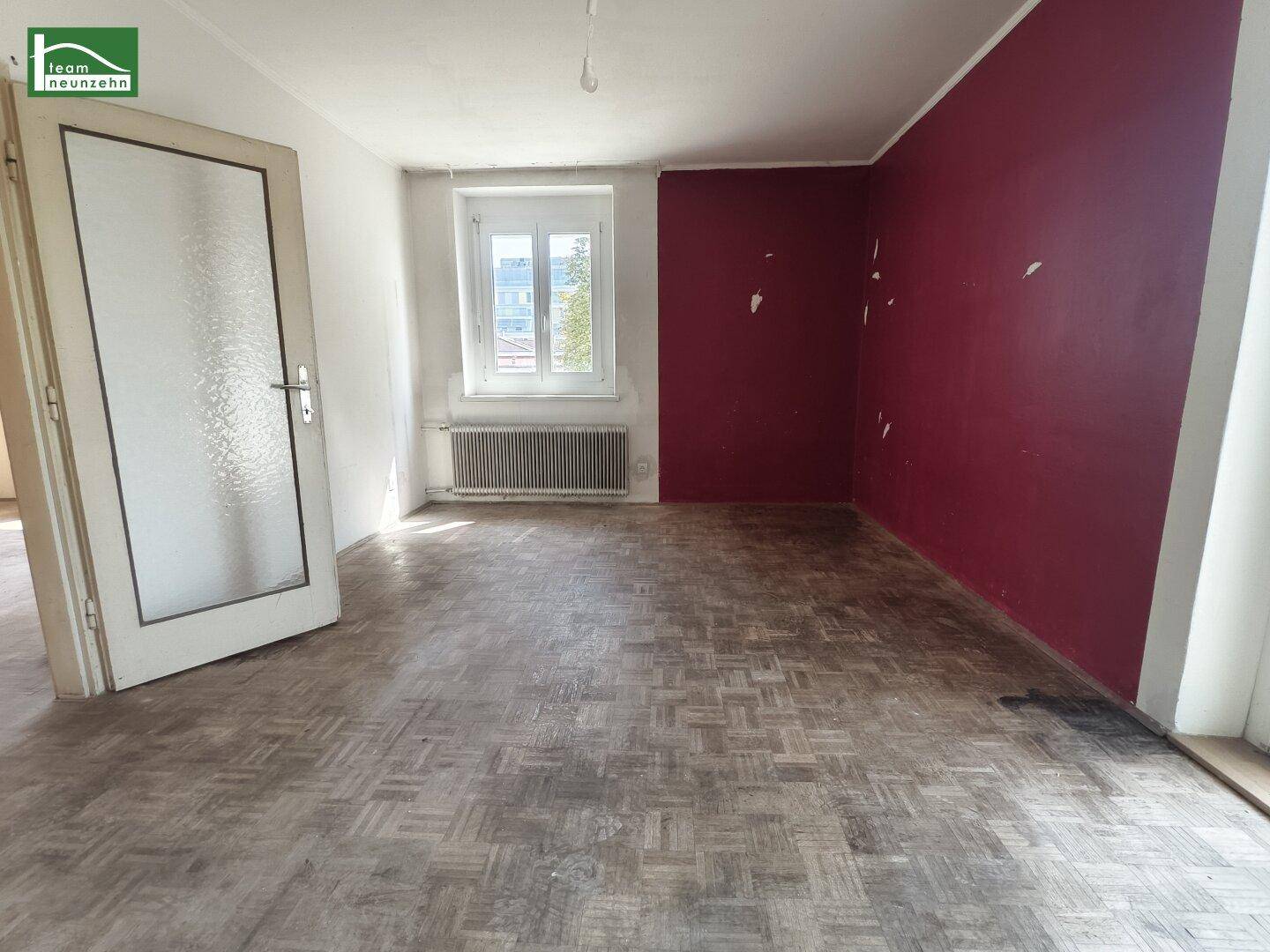 Leeres Zimmer mit abgenutztem Parkettboden, roter Akzentwand und Fenster.