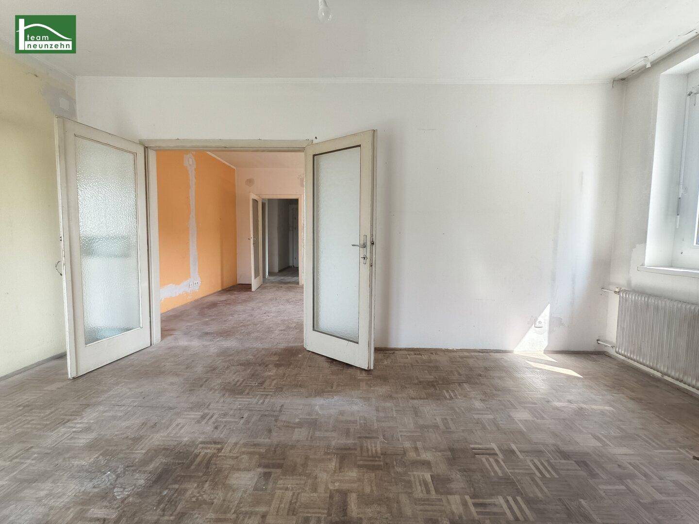 Helles Zimmer mit abgenutztem Parkettboden und Doppeltüren, die zu weiteren Räumen führen.