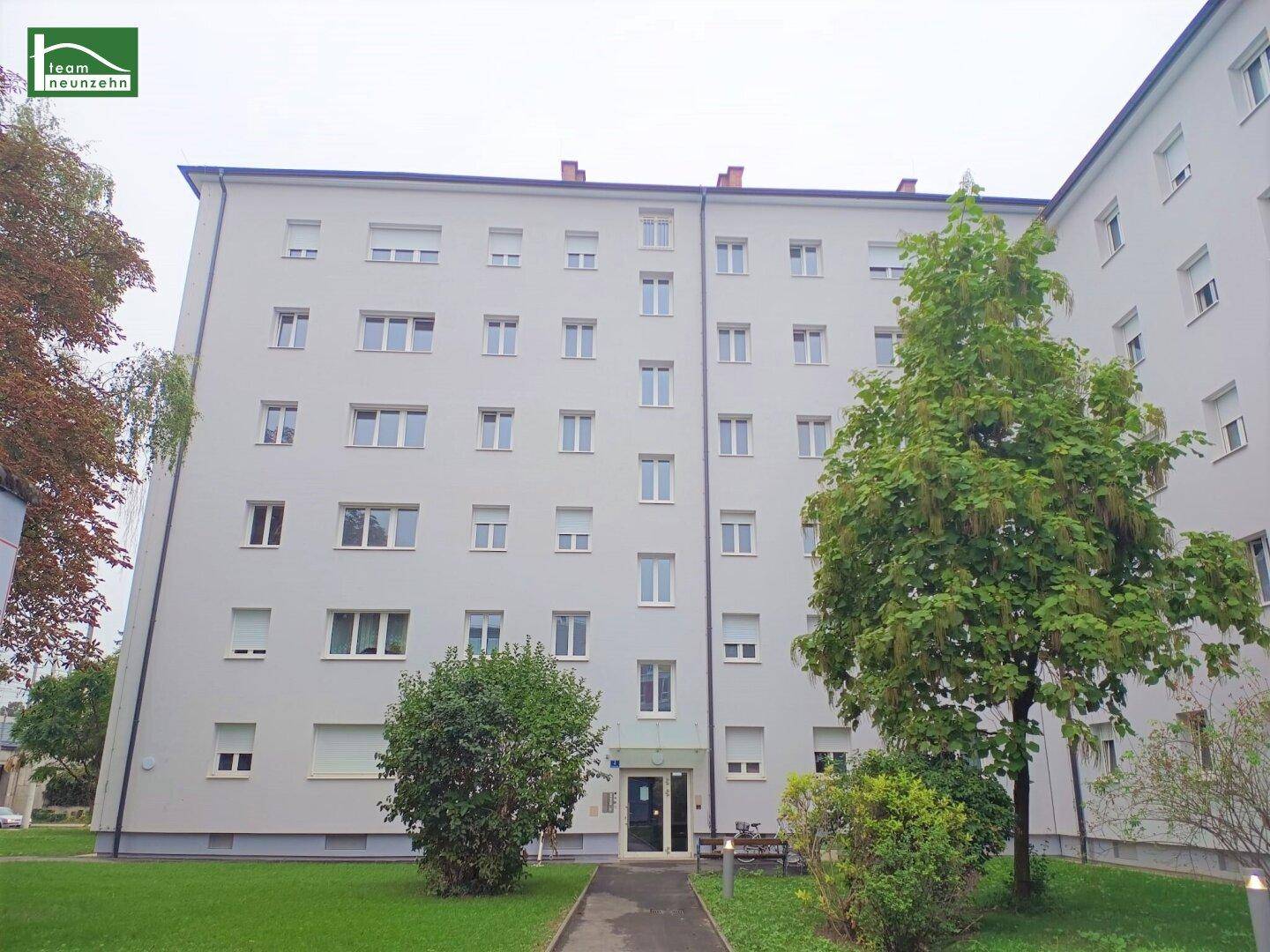 Mehrfamilienhaus mit weißer Fassade, vielen Fenstern und gepflegter Grünanlage.
