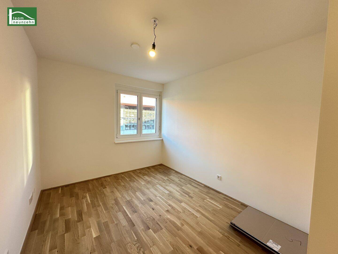 Leeres Zimmer mit Parkettboden und einem Fenster, das viel Tageslicht hereinlässt.