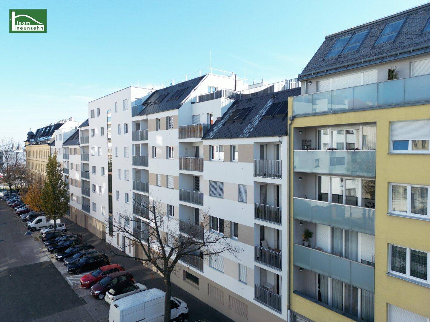 Mehrere moderne Wohngebäude entlang einer Straße mit geparkten Autos und Bäumen.