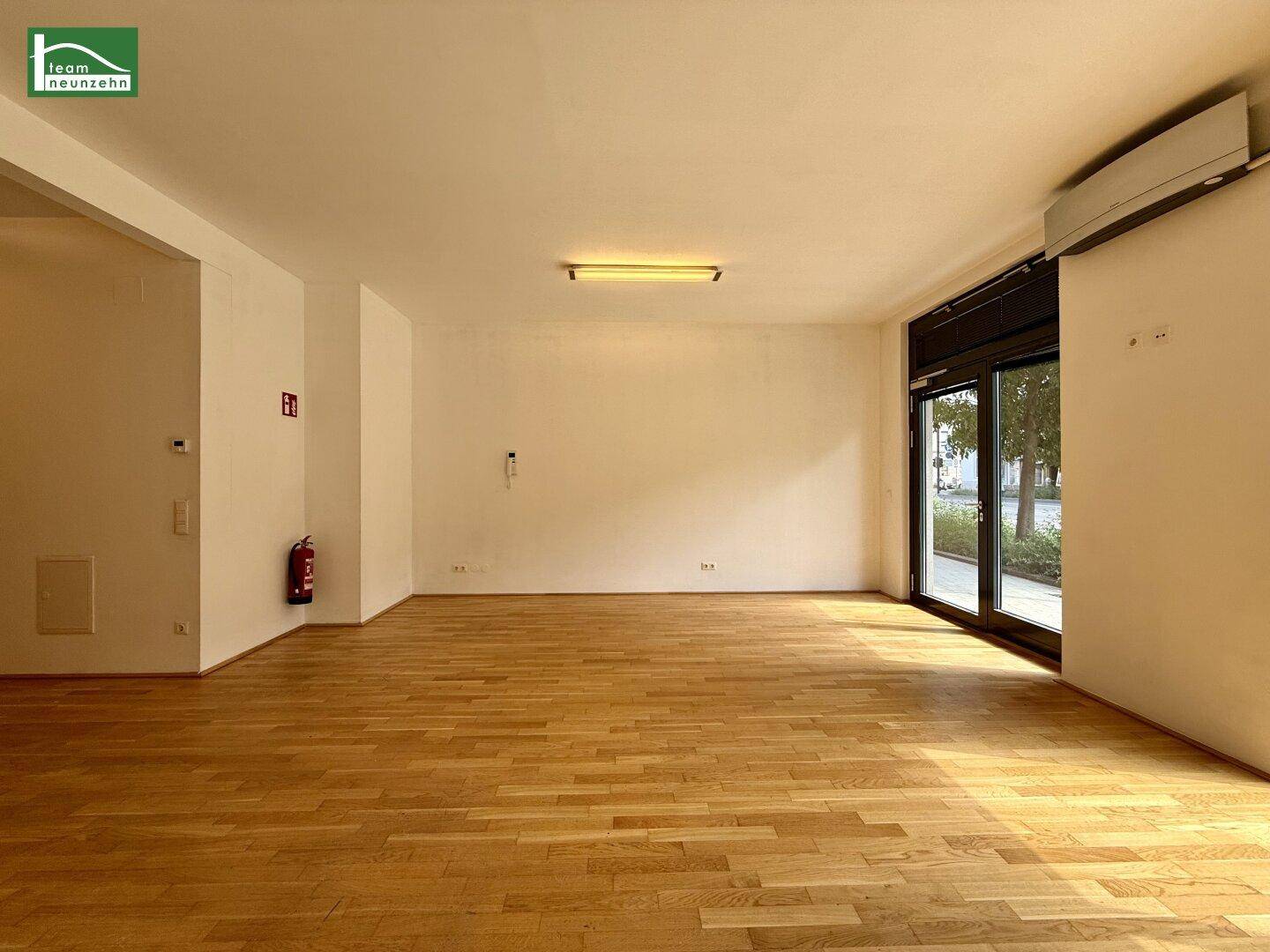 Geräumiger Gewerberaum mit Holzboden, großen Fenstern zur Straße und Klimaanlage.
