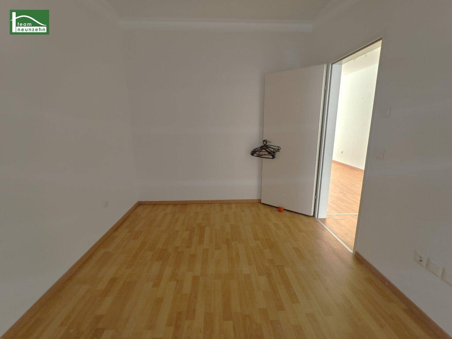 Leeres Zimmer mit Holzboden und weiß gestrichenen Wänden, mit offener Tür zu einem weiteren Raum.
