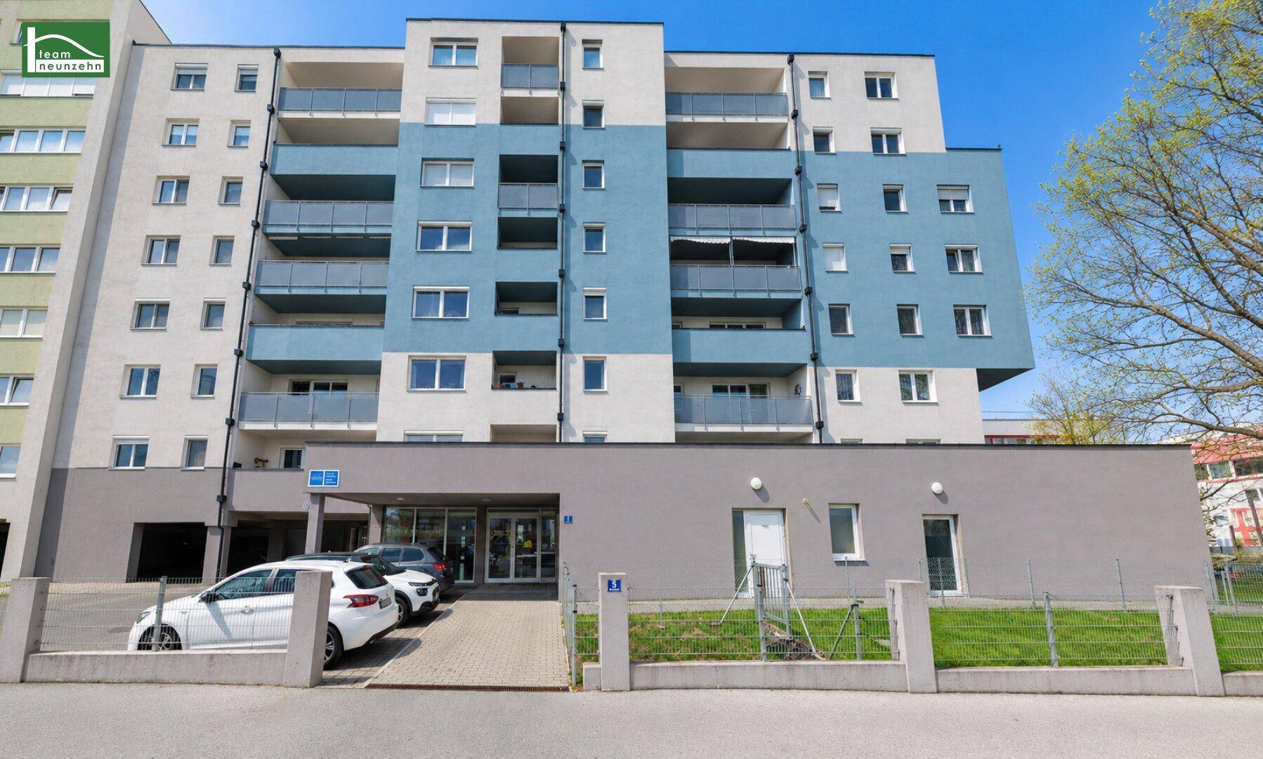 Eingangsbereich eines modernen Wohngebäudes mit Parkplätzen davor und gepflegter Grünfläche.