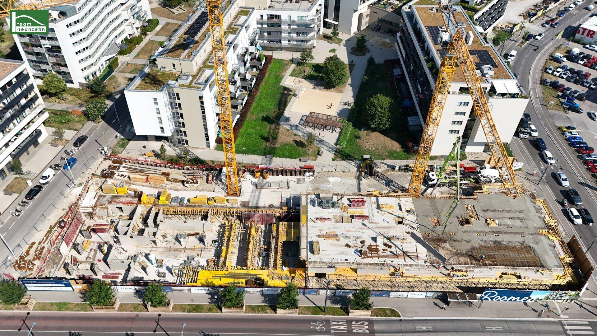 Luftaufnahme einer Baustelle mit Kränen, umgeben von modernen Wohngebäuden und einer belebten Straße.