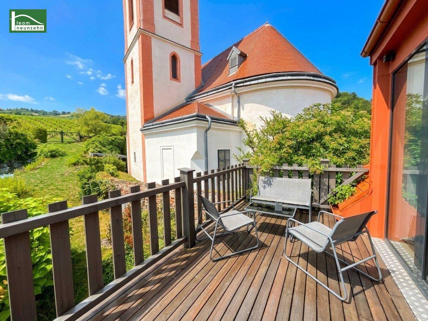 Holzterrasse mit Sitzgelegenheiten und malerischem Blick auf eine Kirche und Weinberge.