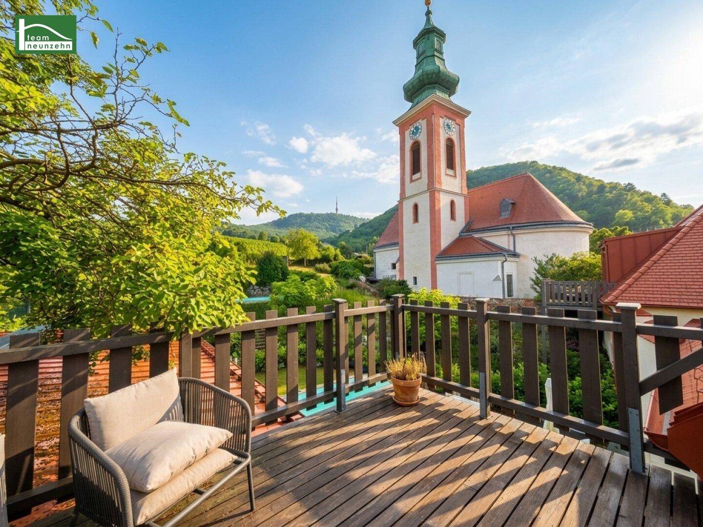 Gemütlicher Holzbalkon mit Sitzgelegenheiten und malerischem Blick auf eine Kirche und die Landschaft.