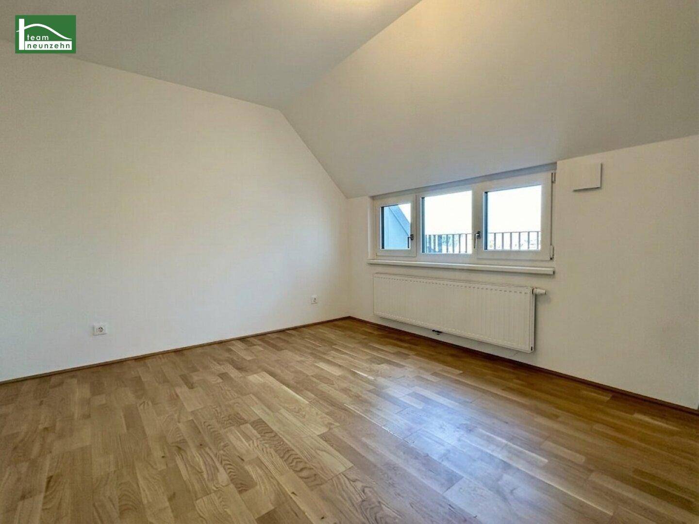 Heller, leerer Raum mit Holzboden, Dachschräge und Fenster mit Blick nach draußen.