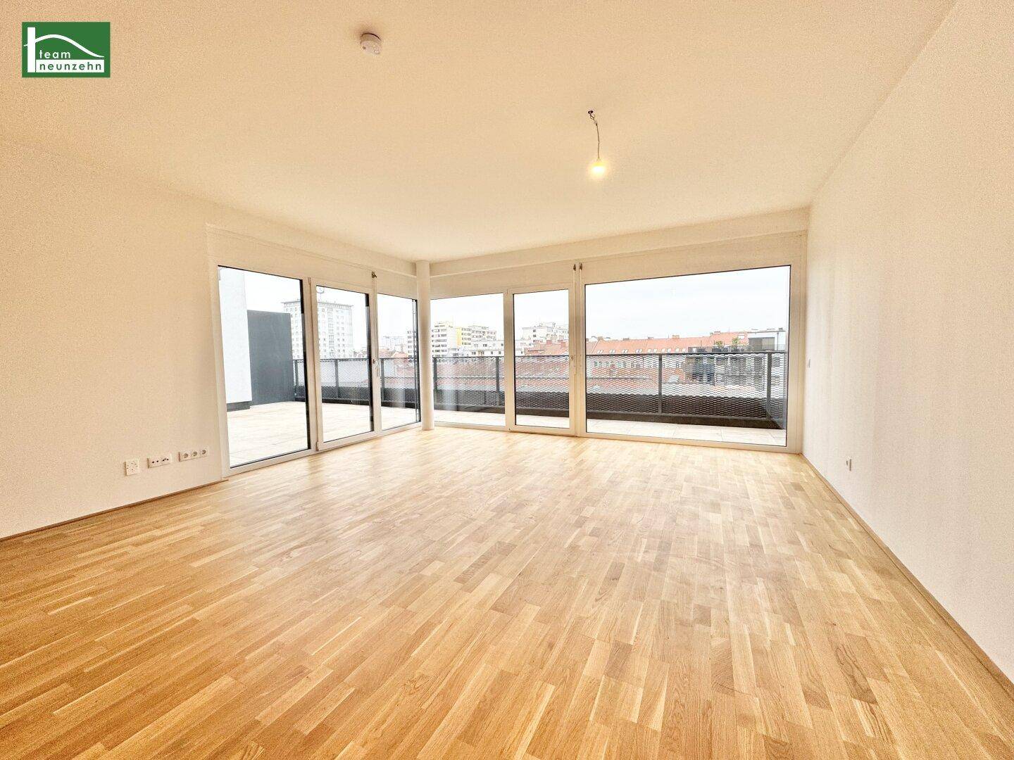 Geräumiges, leeres Wohnzimmer mit Holzboden und bodentiefen Fenstern, die viel Licht hereinlassen.