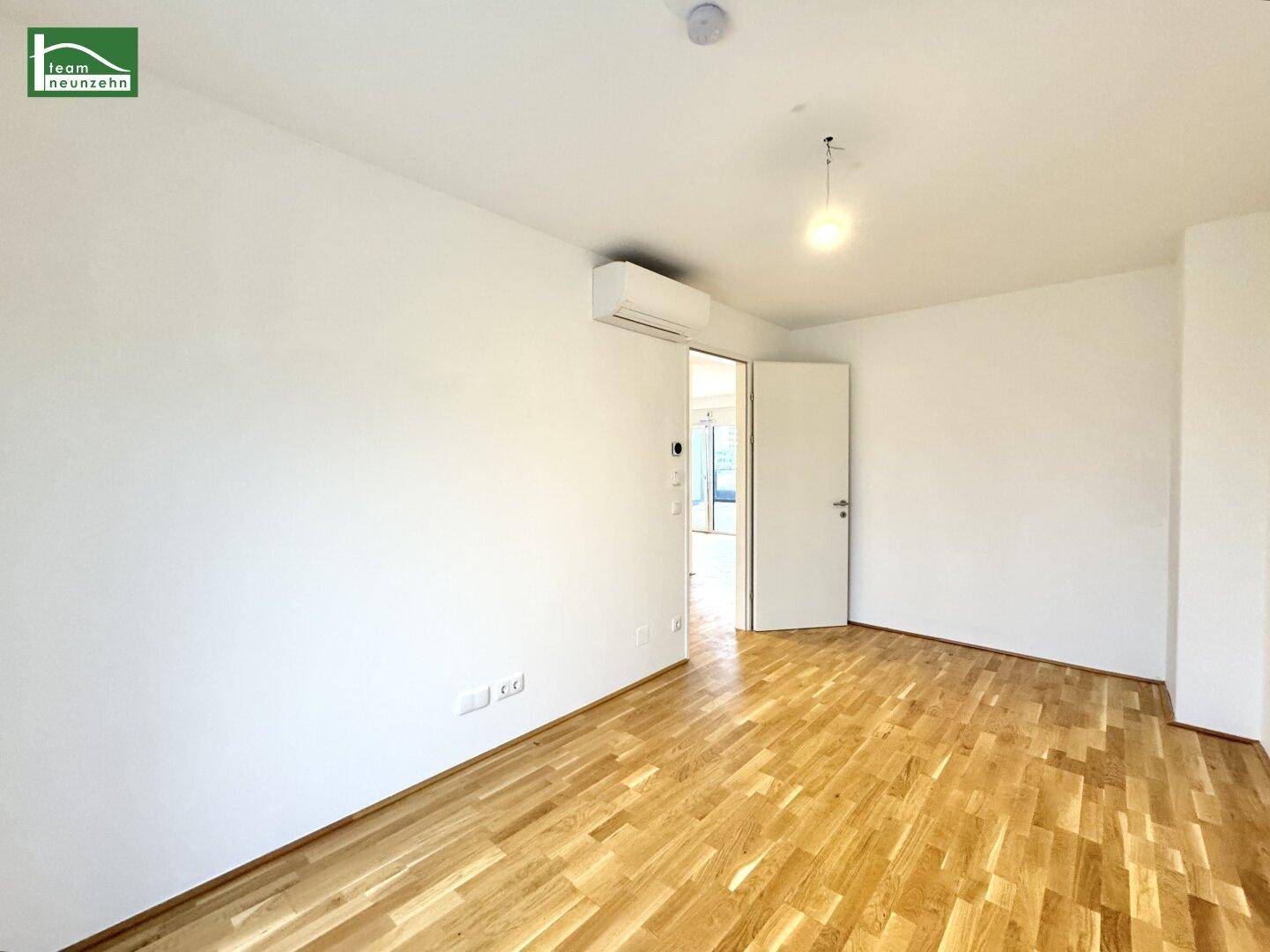 Leeres Zimmer mit Parkettboden, Klimaanlage und Zugang zu weiteren Räumen.