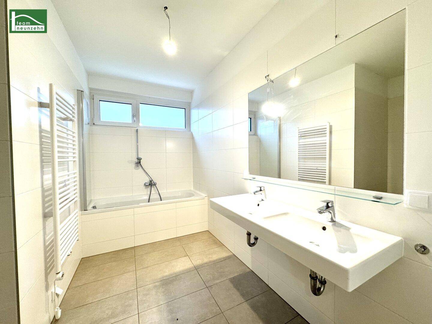 Helles Badezimmer mit Doppelwaschbecken, Badewanne, beheiztem Handtuchhalter und Fenster.