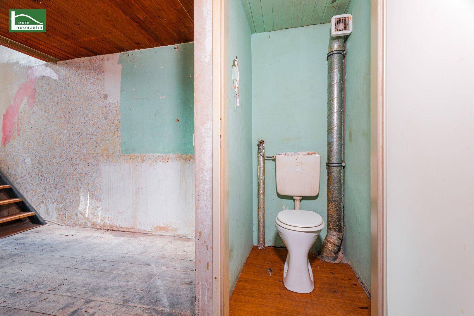 Einfache Toilette in einem kleinen Raum mit Holzdecke und sichtbaren Rohren.