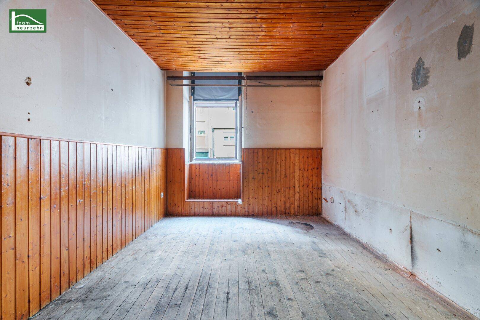 Leerer Raum mit Holzdecke, Dielenboden und einem Fenster, das auf einen Innenhof blickt.