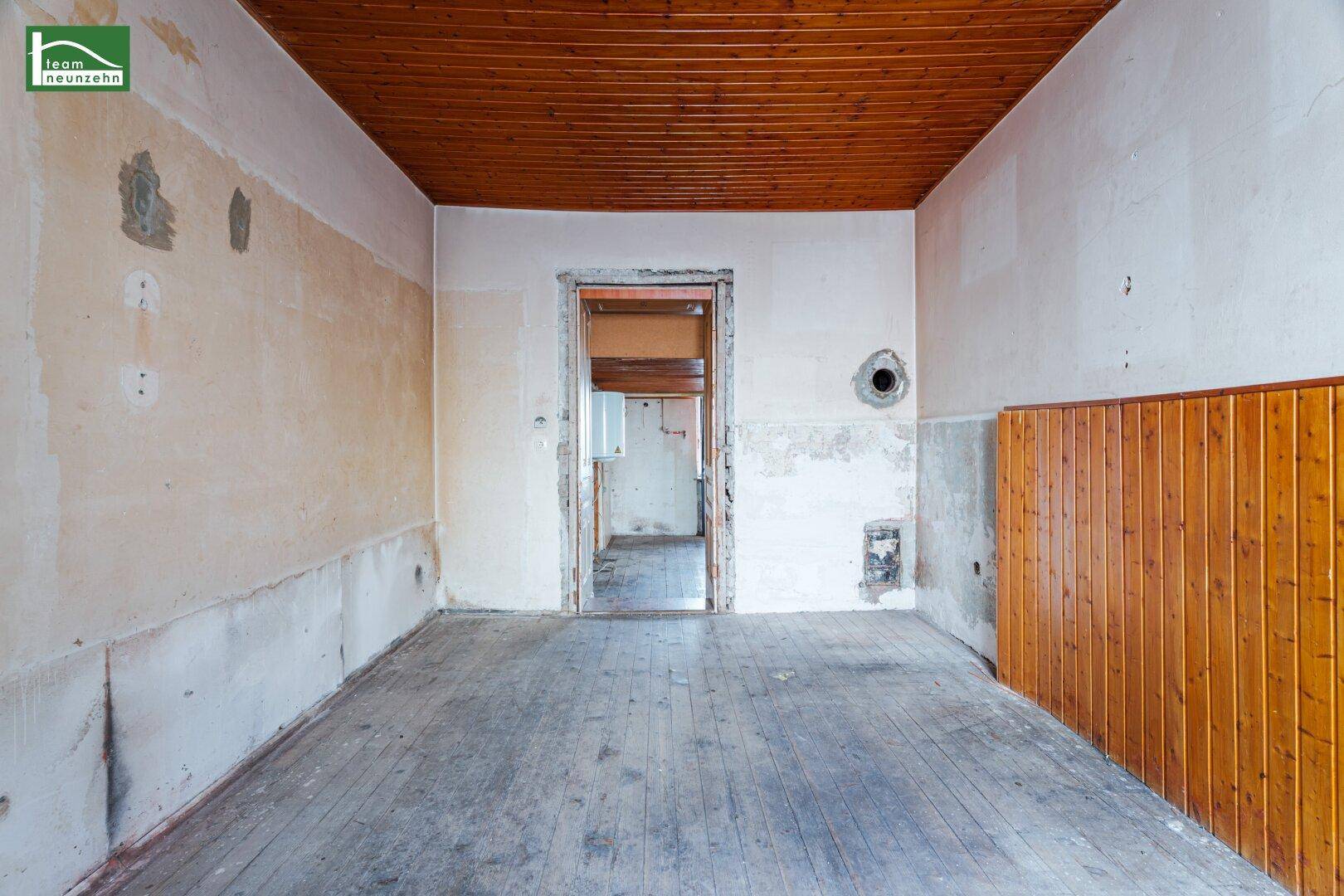 Renovierungsbedürftiger Raum mit Holzboden, Holzdecke und teilweise verputzten Wänden.