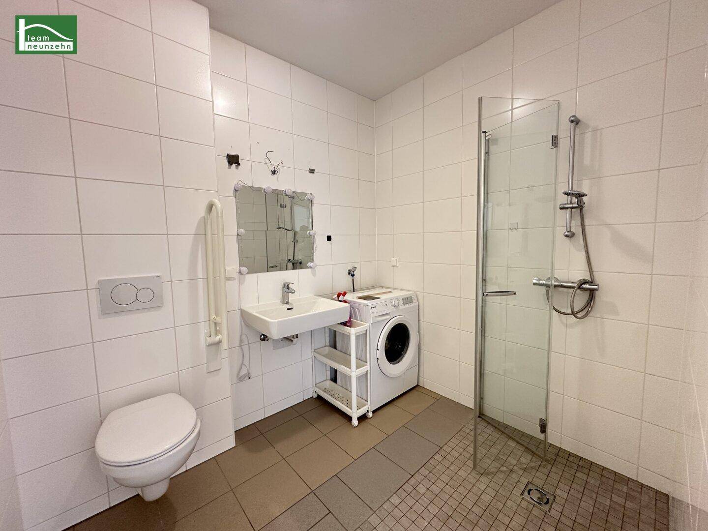 Modernes Badezimmer mit ebenerdiger Dusche, wandhängendem WC und Waschmaschinenanschluss, hell gefliest.