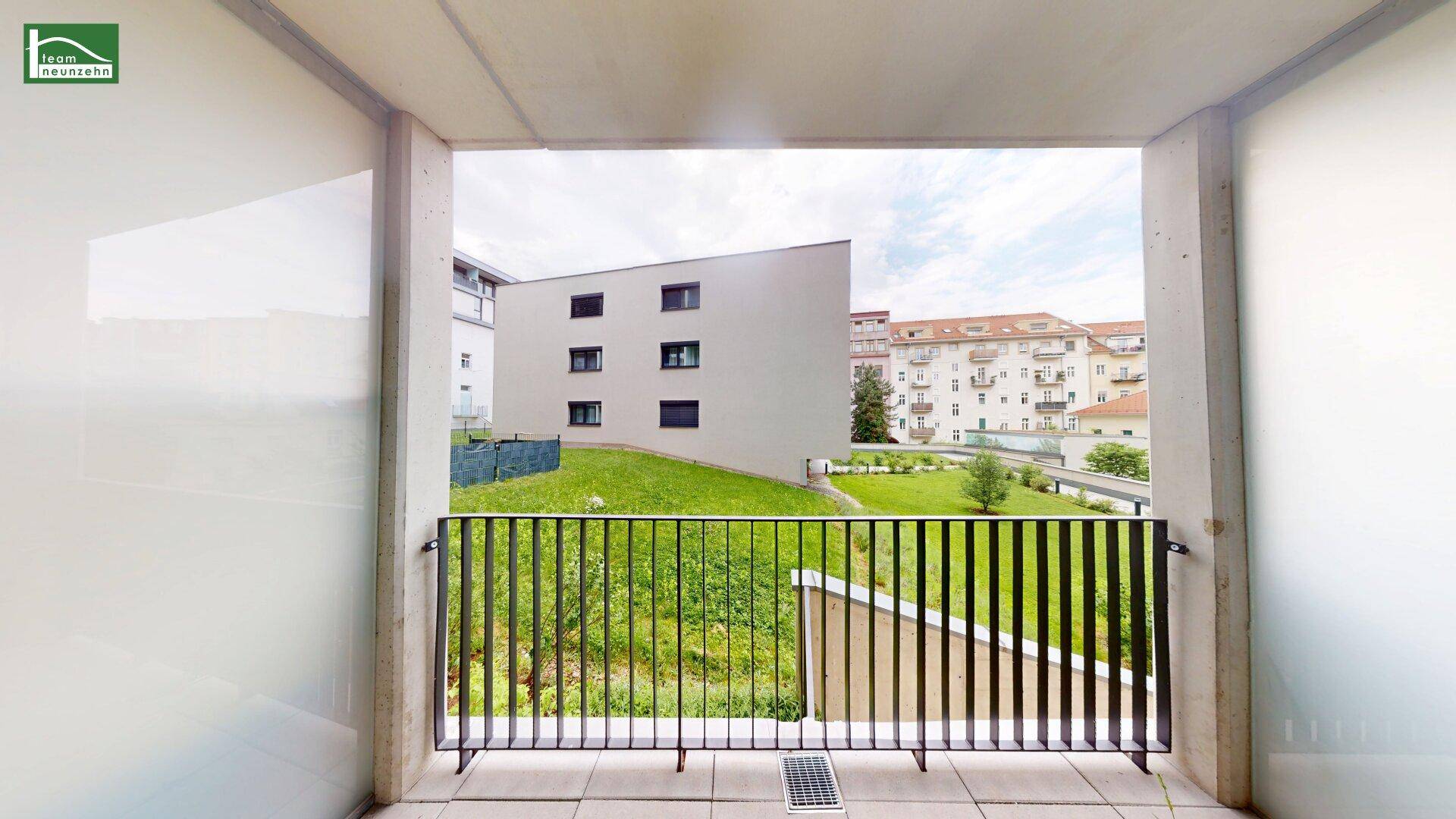 Balkon mit Metallgeländer und Blick auf eine grüne Rasenfläche und umliegende Gebäude.