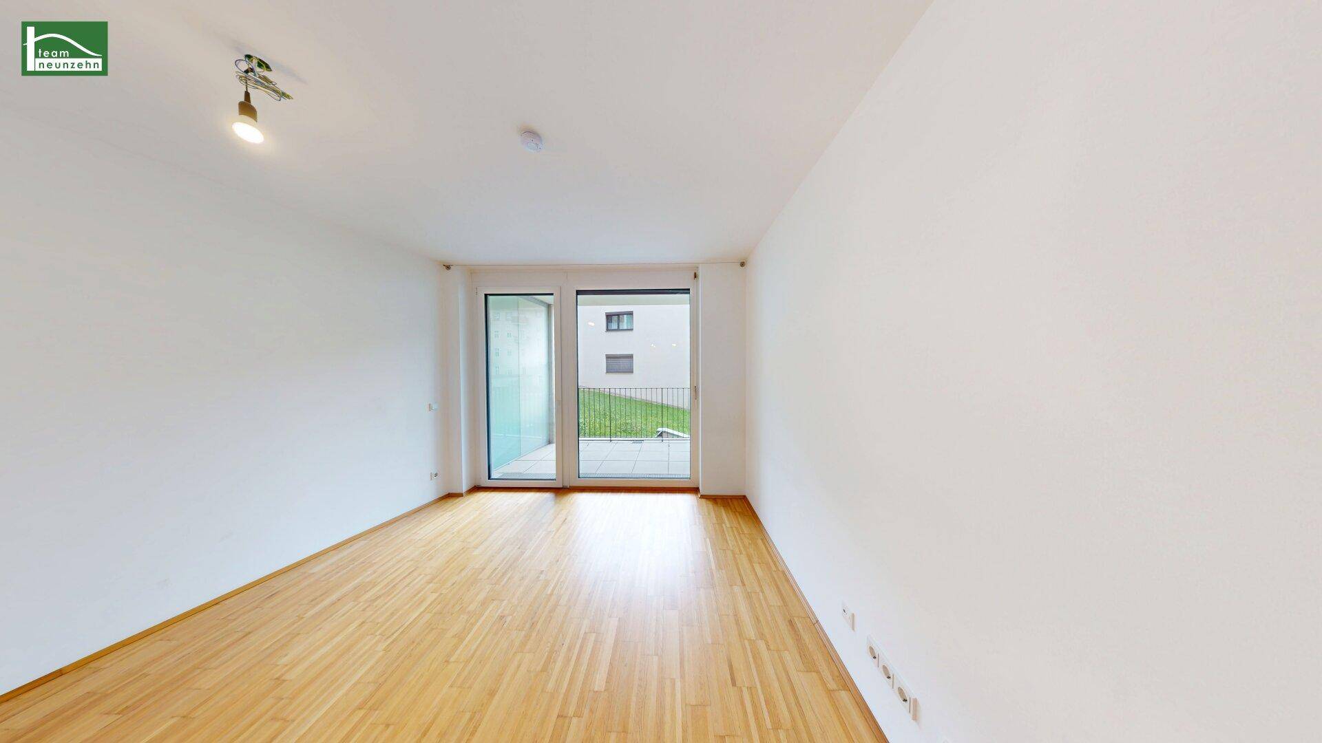 Leerer, heller Wohnraum mit Holzboden und großen Fenstern zum Außenbereich.